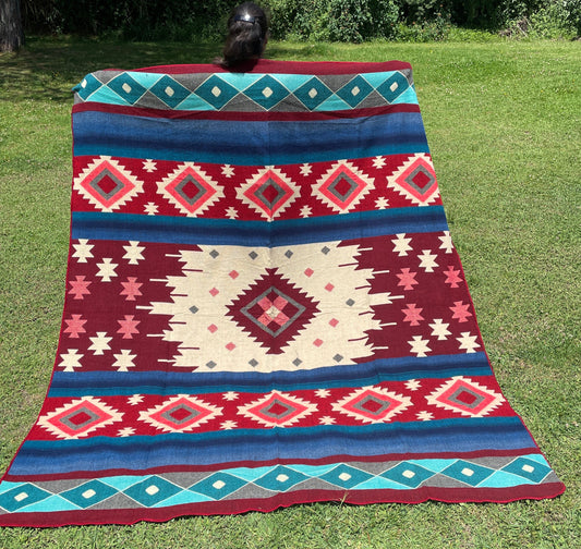 TomHats alpaca wool blanket with traditional Navajo patterns in red and blue on a green meadow.