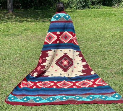 TomHats alpaca wool blanket with traditional Navajo patterns in red and blue on a green meadow.