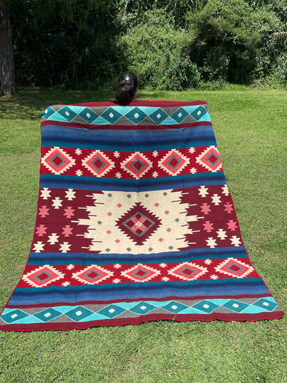 TomHats alpaca wool blanket with traditional Navajo patterns in red and blue on a green meadow.