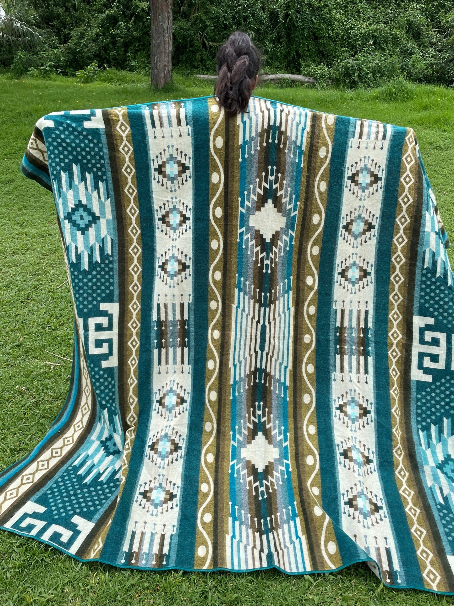 TomHats alpaca blanket in turquoise with traditional indigenous patterns, draped over a person outdoors.