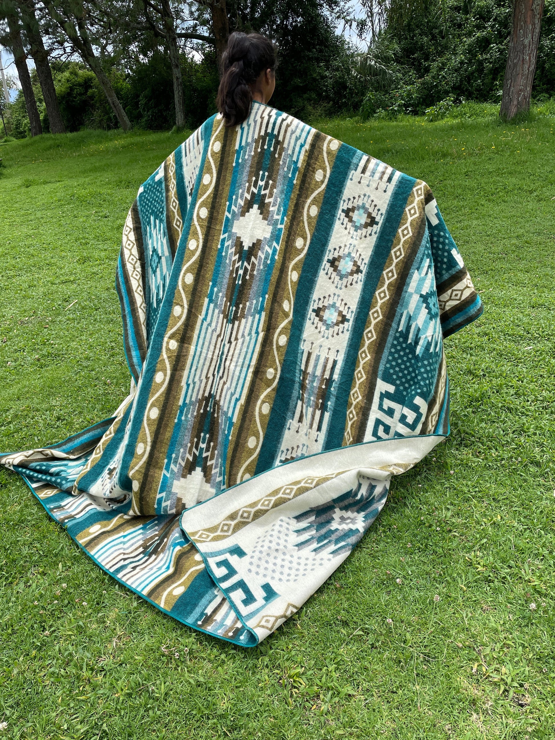 TomHats alpaca blanket in turquoise with traditional indigenous patterns, draped over a person outdoors.