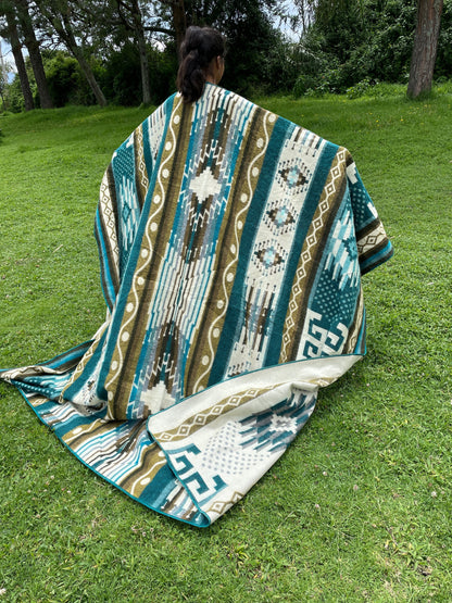 TomHats alpaca blanket in turquoise with traditional indigenous patterns, draped over a person outdoors.