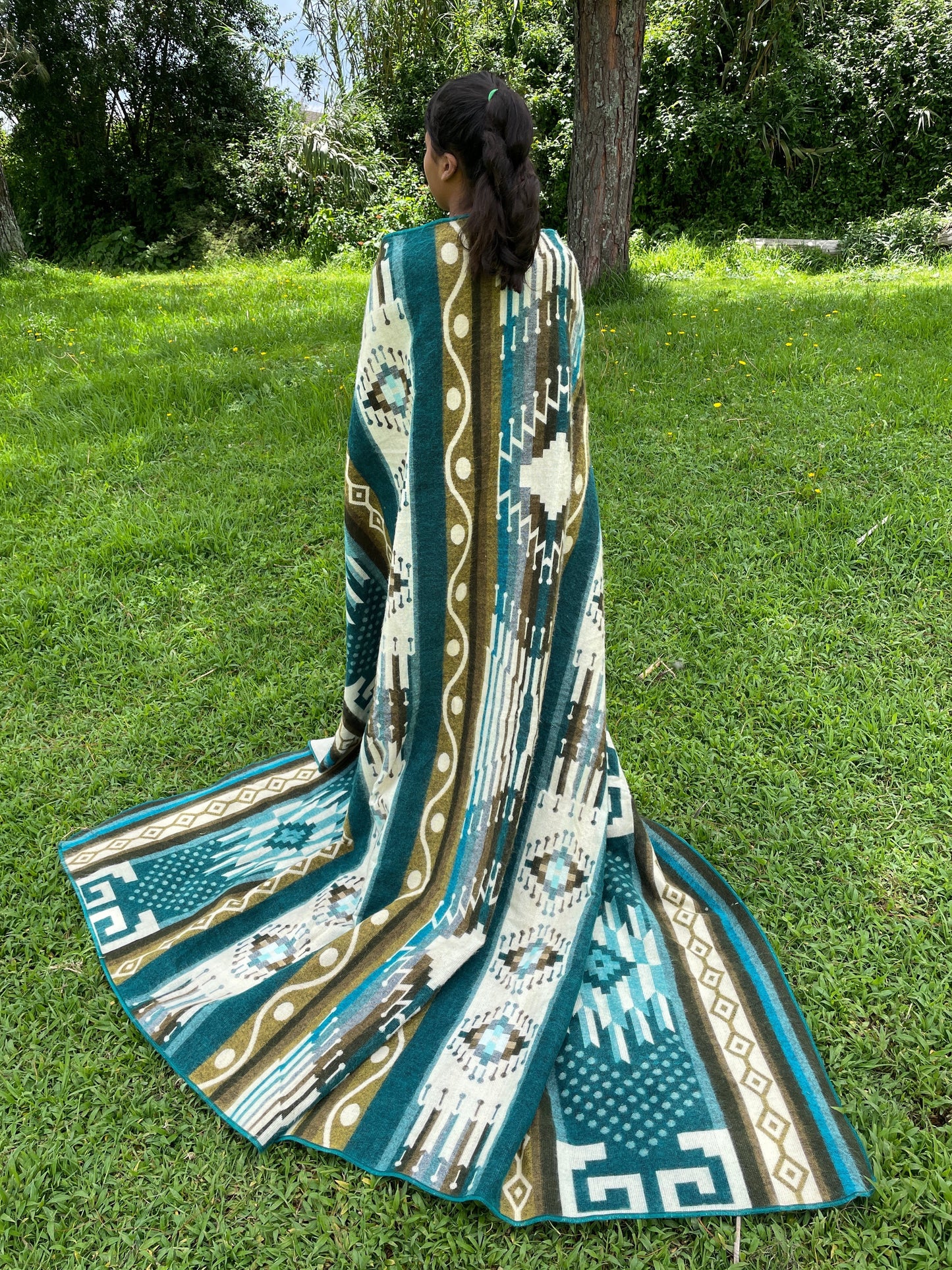 TomHats alpaca blanket in turquoise with traditional indigenous patterns, draped over a person outdoors.
