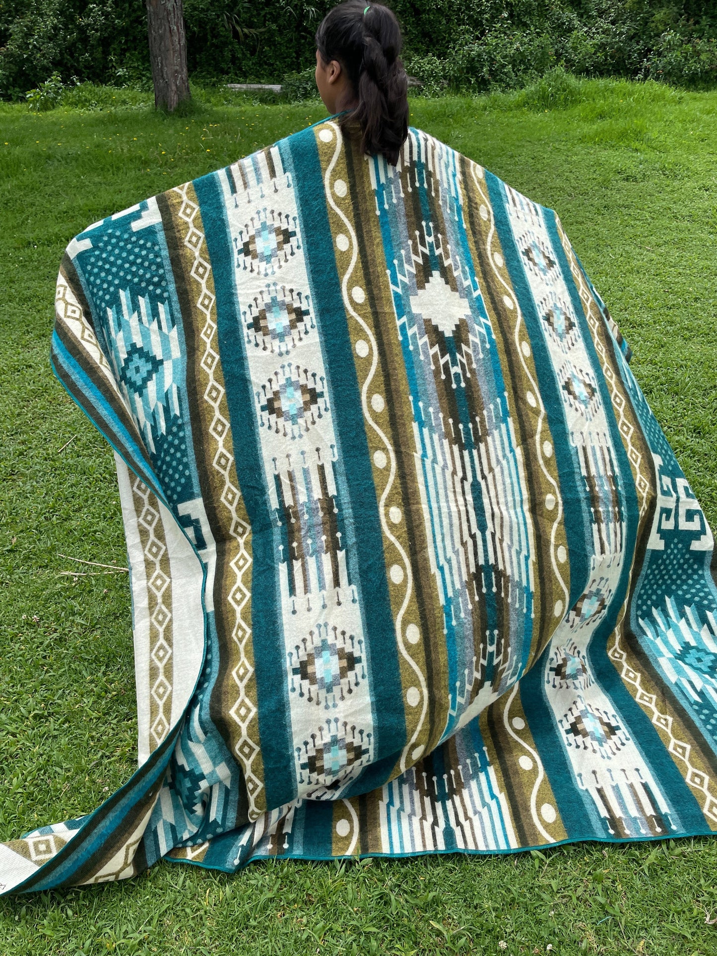 TomHats alpaca blanket in turquoise with traditional indigenous patterns, draped over a person outdoors.