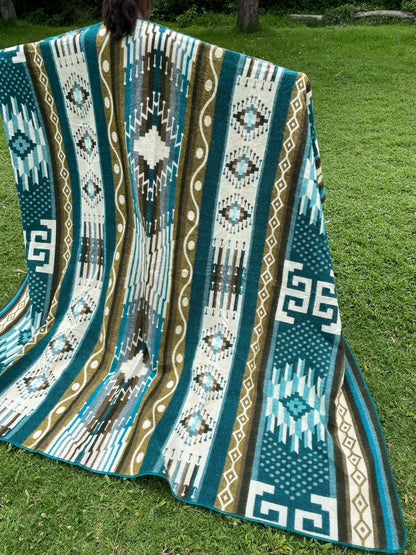 TomHats alpaca blanket in turquoise with traditional indigenous patterns, draped over a person outdoors.