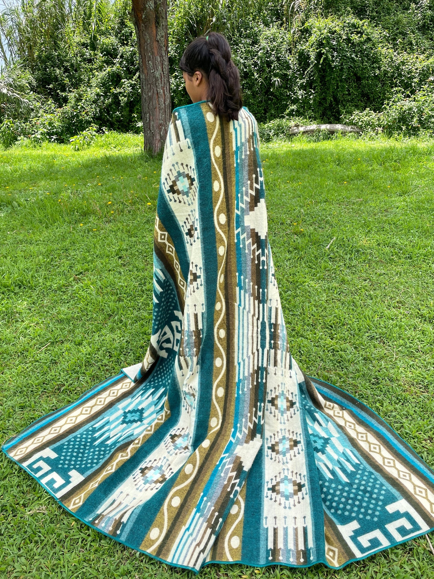 TomHats alpaca blanket in turquoise with traditional indigenous patterns, draped over a person outdoors.