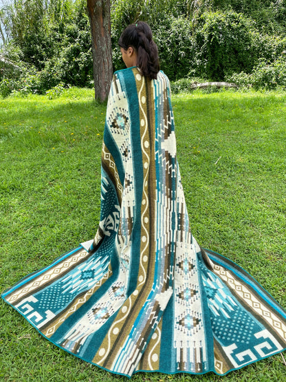 TomHats alpaca blanket in turquoise with traditional indigenous patterns, draped over a person outdoors.